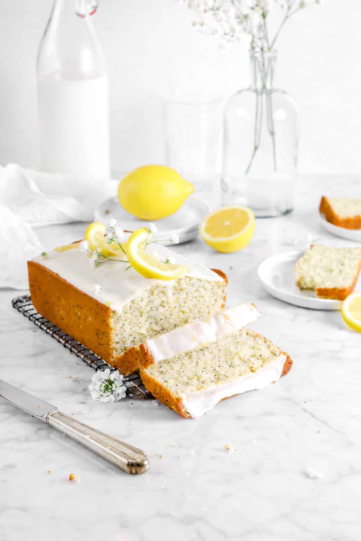 lemon poppy seed bread on wire cooling tack with two slices laying in front, two slices behind, lemons behind, a vase, white napkin, and jug of milk