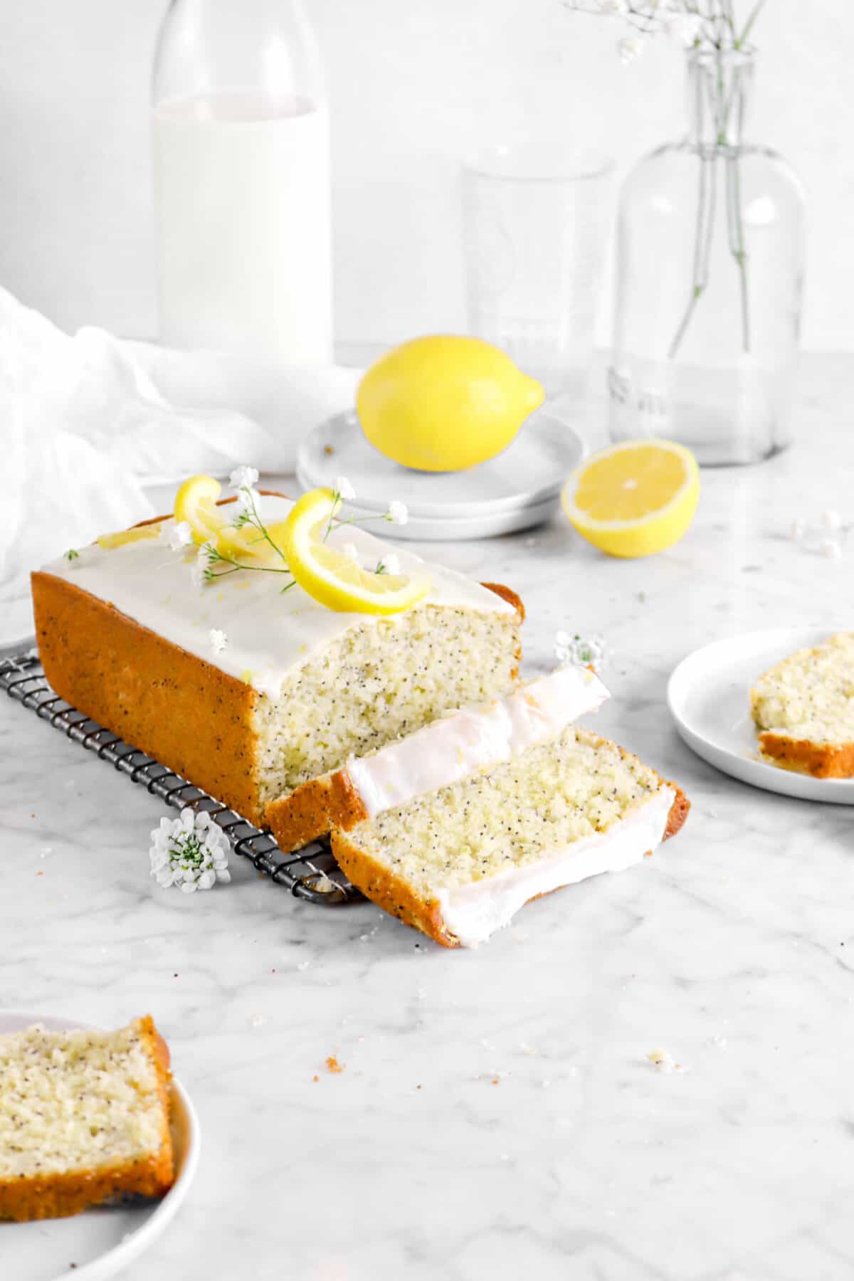 lemon poppy seed bread on wire cooling rack with two slices laying in front with two slices on white plates with lemon behind, jug of milk, white napkin, and vase