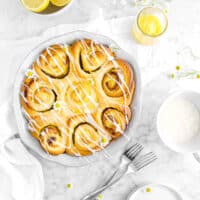 overhead shot of lemon rolls in pie pan with a napkin, forks, lemons, flowers, lemon curd, and plates