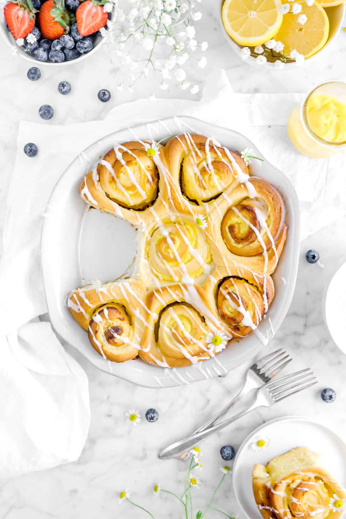 overhead shot of lemon sweet rolls with one taken out of the pan with berries, a white napkin, lemon curd, and forks