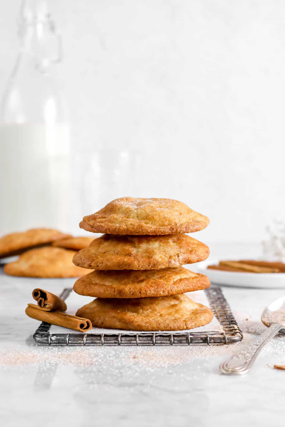 four snickerdoodle cookies stacked on wire cooling rack with two cinnamon sticks, a spoon, more cookies behind, and glass of milk