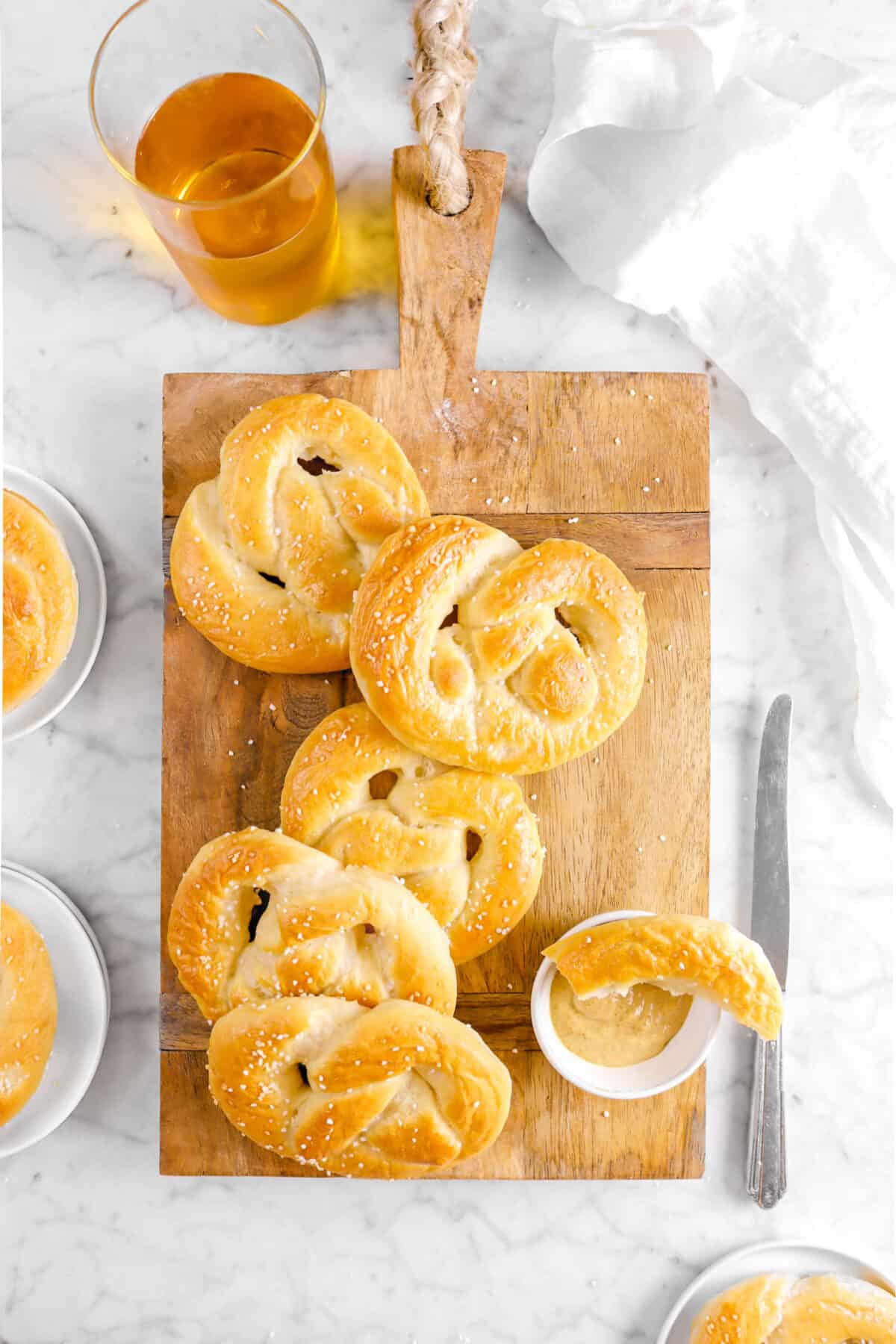 overhead shot of pretzels on wood board with pretzel in mustard bowl, a glass of beer, a knife, and white napkin