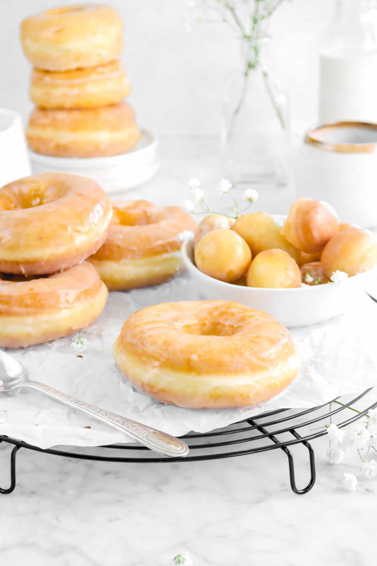 glazed doughnuts on wire cooling rack with doughnut holes in a white bowl, flowers, a stack of doughnuts behind