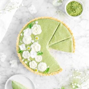 matcha tart with piece missing on a white plate with mint leaves and fresh flowers around