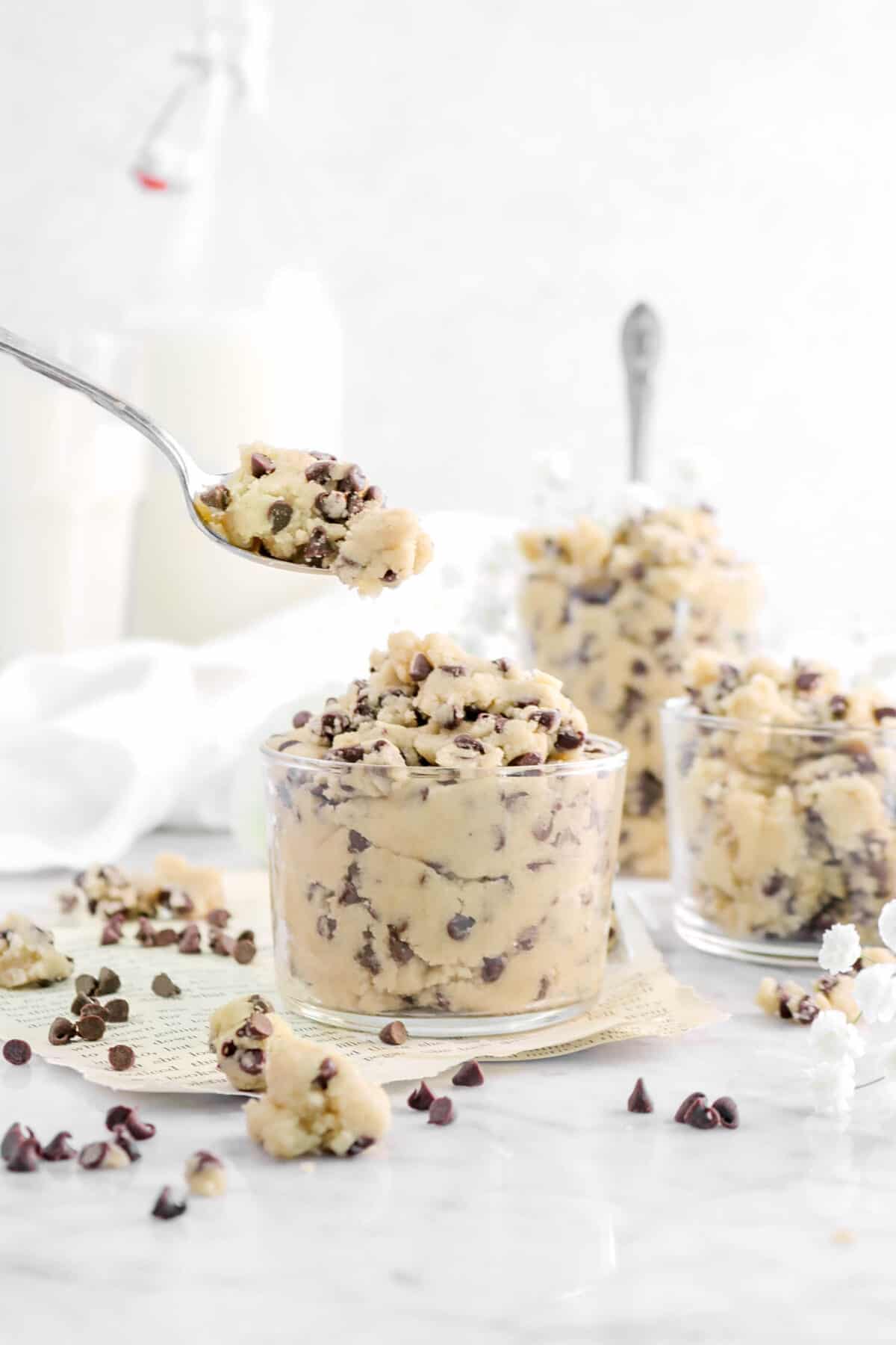 spoonful of cookie dough being held over glass with more cookie dough behind and glasses of milk