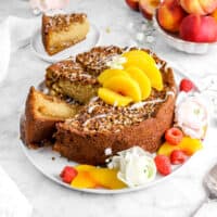 peach coffee cake on white plate with two slices laying beside cake, one behind on white plate, with fresh berries, peach slices, and flowers