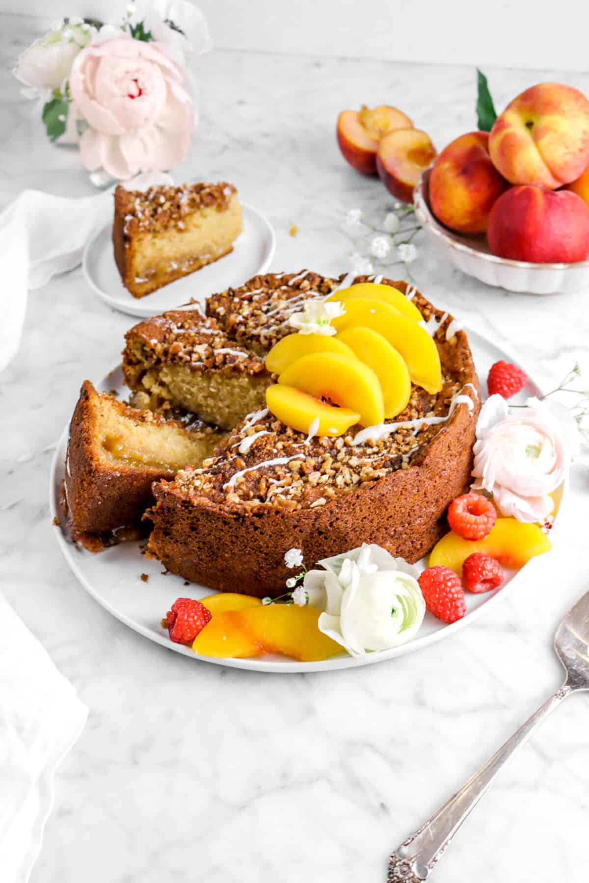 peach coffee cake on white plate with two slices laying beside cake, one behind on white plate, with fresh berries, peach slices, and flowers