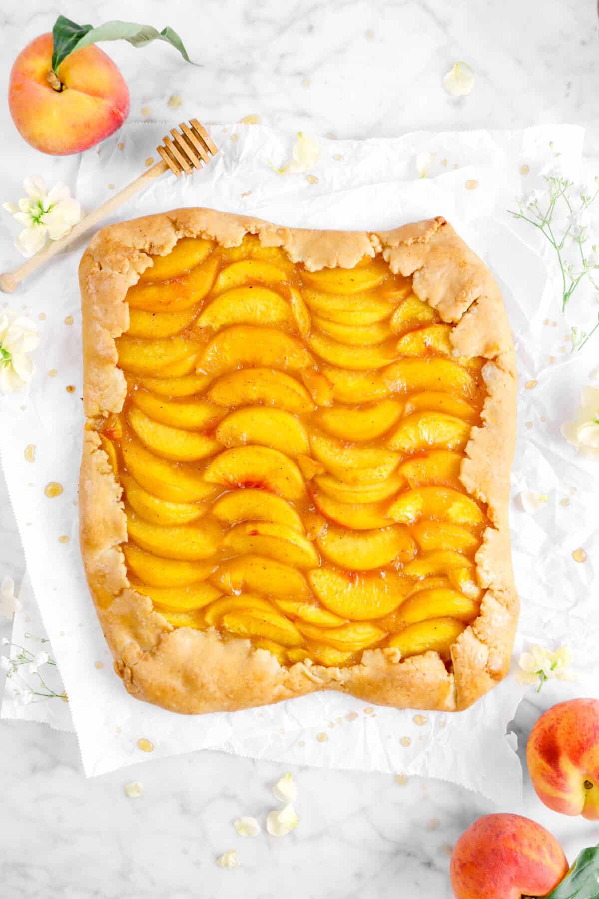 peach galette on two pieces of parchment paper, fresh peaches, flowers, and a honey dipper