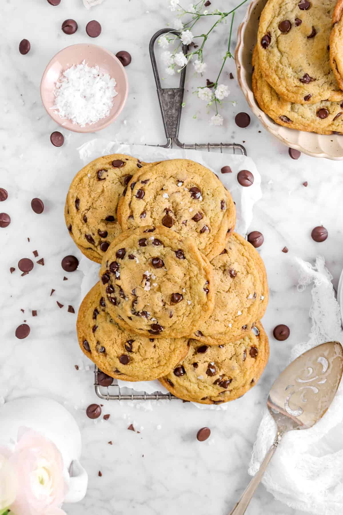 six chocolate chip cookies on serving tray with chocolate chips, a bowl of cookies, a bowl of salt, flowers, and a cake knife