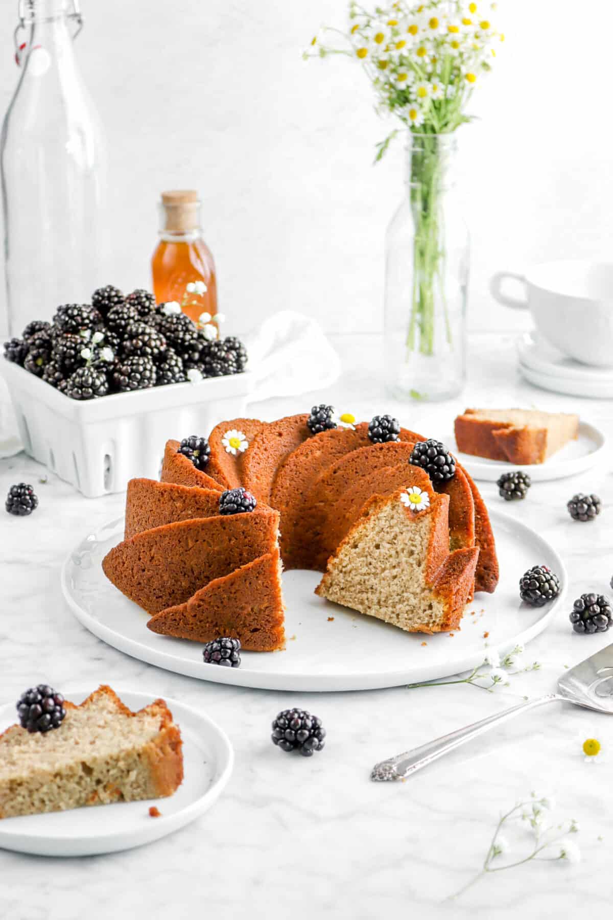 sliced blackberry apple spice cake on white plate with blackberries, flowers, and a jar of honey