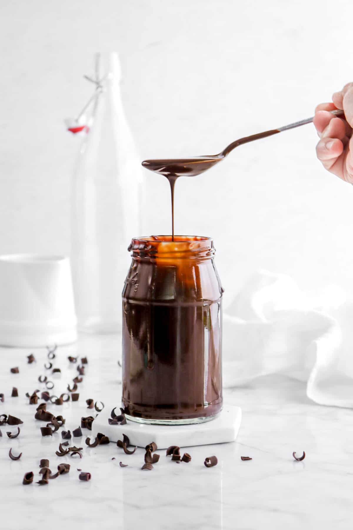 chocolate sauce with a spoon pouring sauce into the jar with chocolate curls, a white napkin, and glasses behind