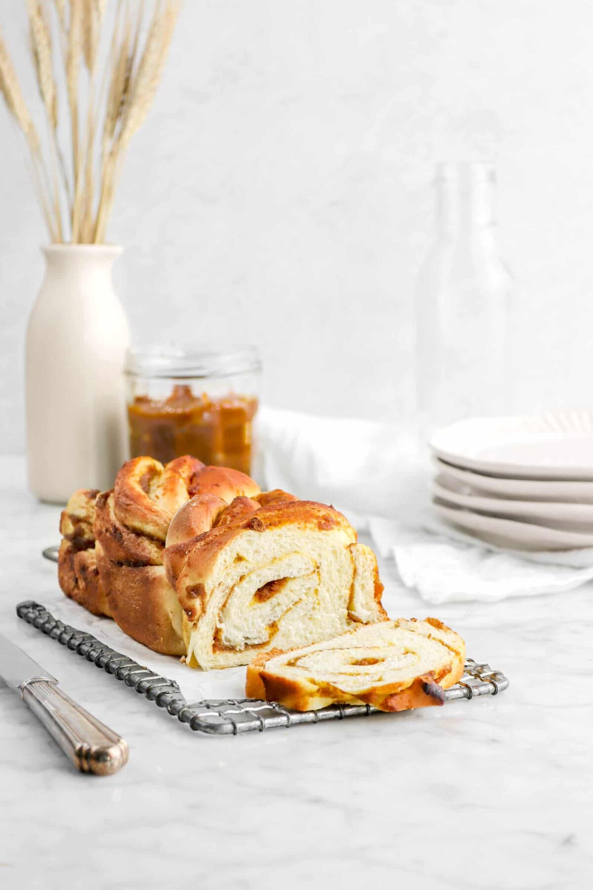 pumpkin butter babka on wire rack with slice laying in front with jar of wheat, a jar of pumpkin butter, and plates behind