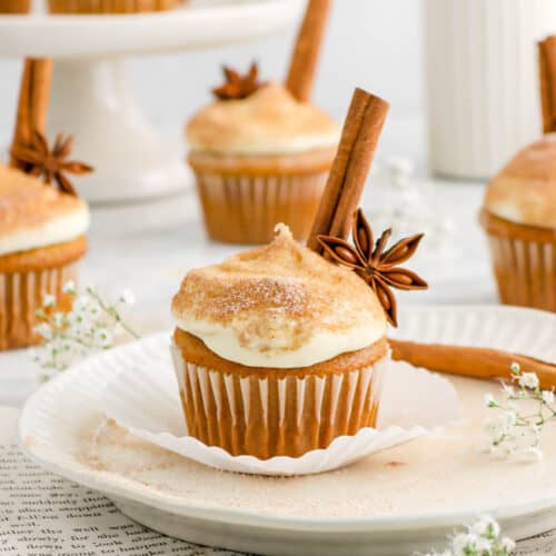pumpkin cupcake with star anise and cinnamon stick on white plate, book pages underneath, more cupcakes behind, and flowers