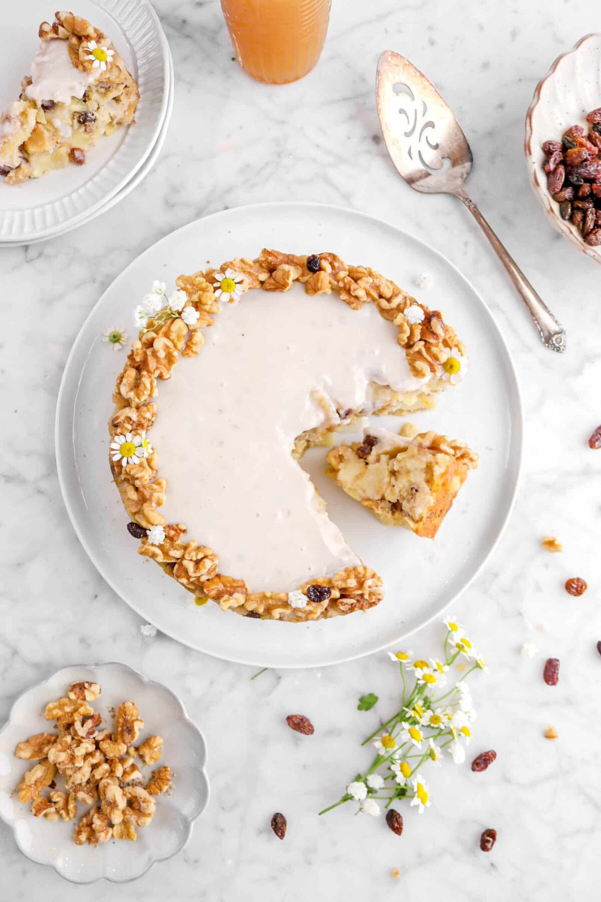 cake with slice on its side, walnuts, raisins, and flowers around white plate