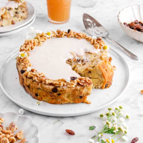 apple breakfast cake on white plate with slice turned on it's side, flowers, walnuts, and raisins around