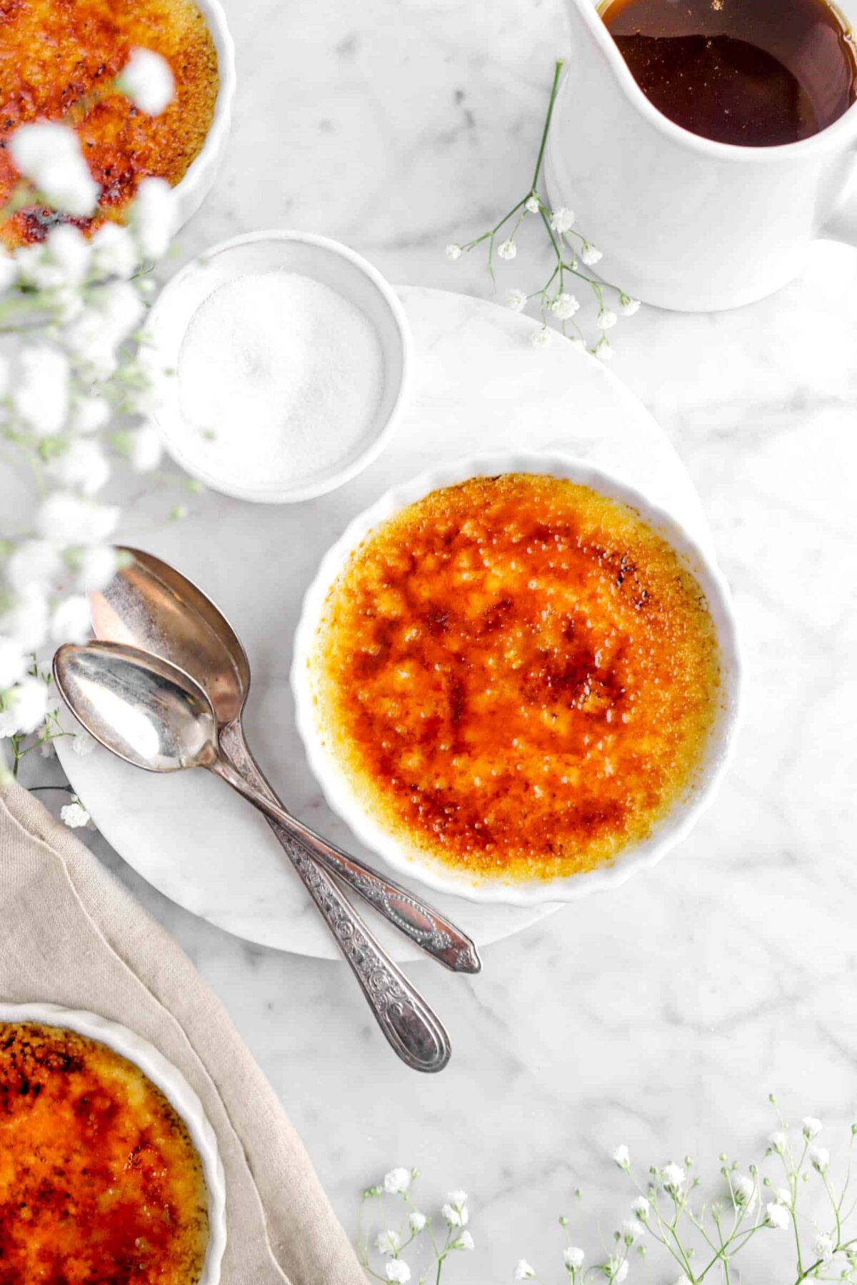 overhead shot of creme brûlée with two spoons, bowl of sugar, and flowers