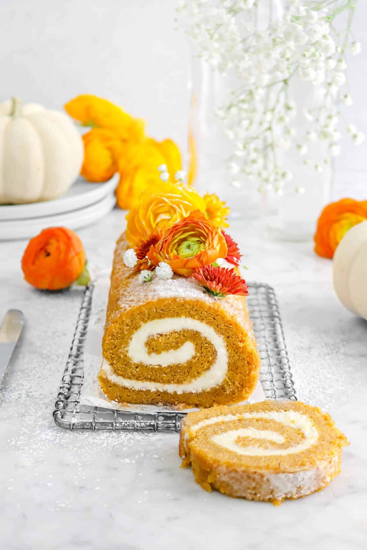 pumpkin roll on wire rack with small piece of parchment, flowers, on top, more flowers around, and two white pumpkins