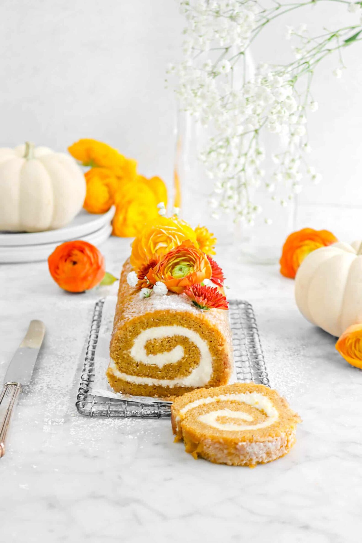 pulled away shot of pumpkin roll with flowers around, a slice laying in front, and two white pumpkins