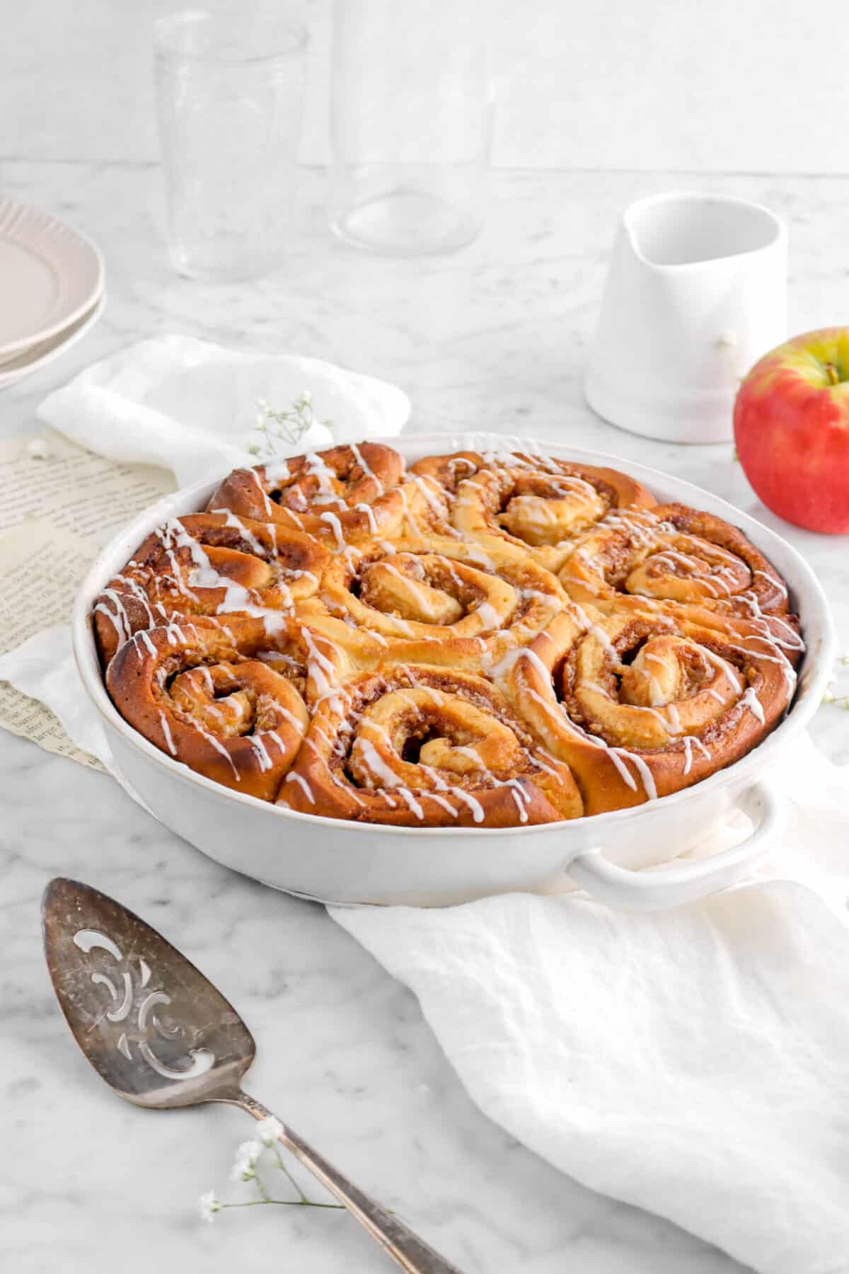 eight apple pie cinnamon rolls in white baking dish with cake knife on white napkin with apple, two glasses behind, and two paltes