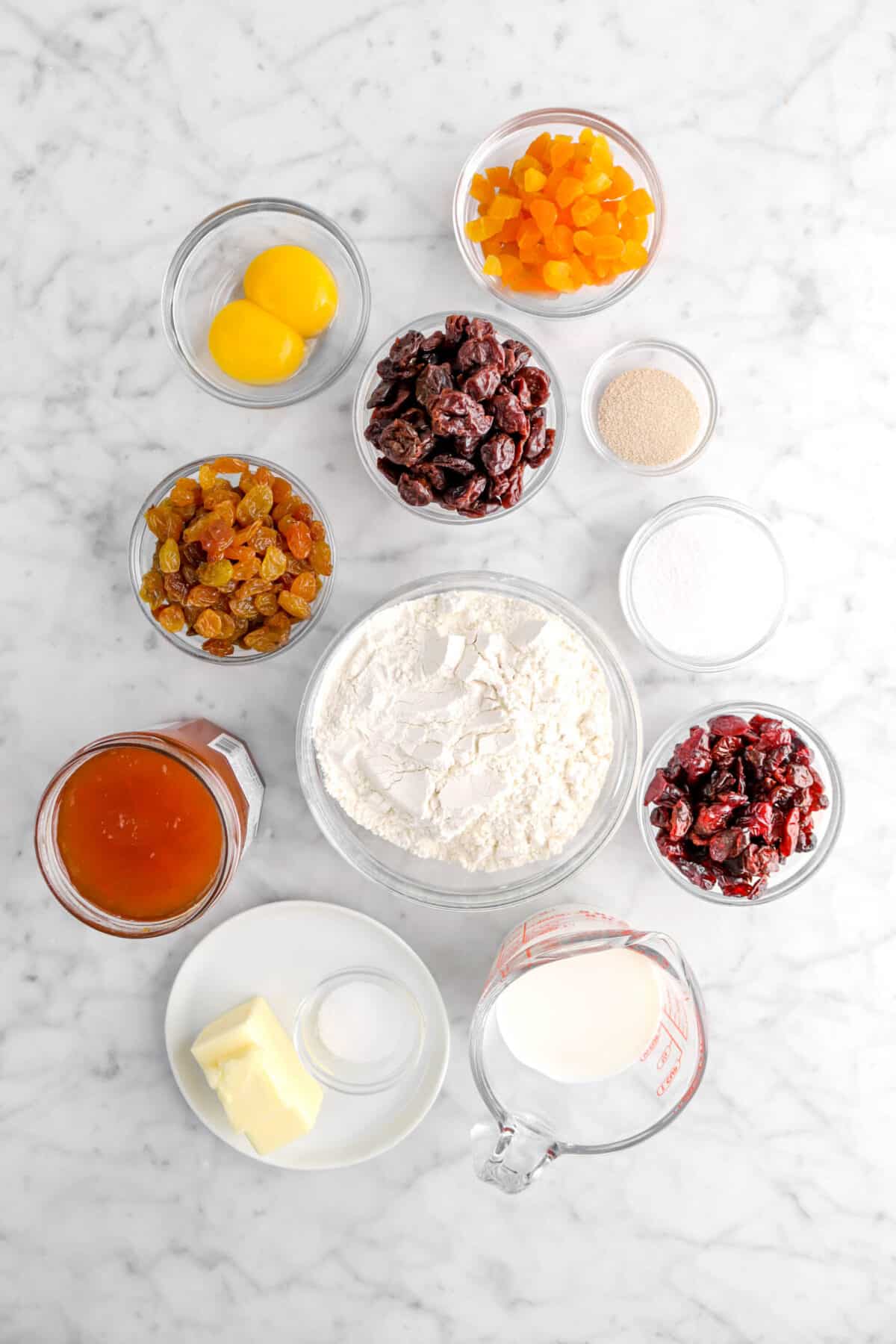 chopped apricot, two egg yolks, dried cranberries, dried golden raisins, yeast, sugar, dried cranberries, flour, apricot jam, butter, salt, and milk on marble counter