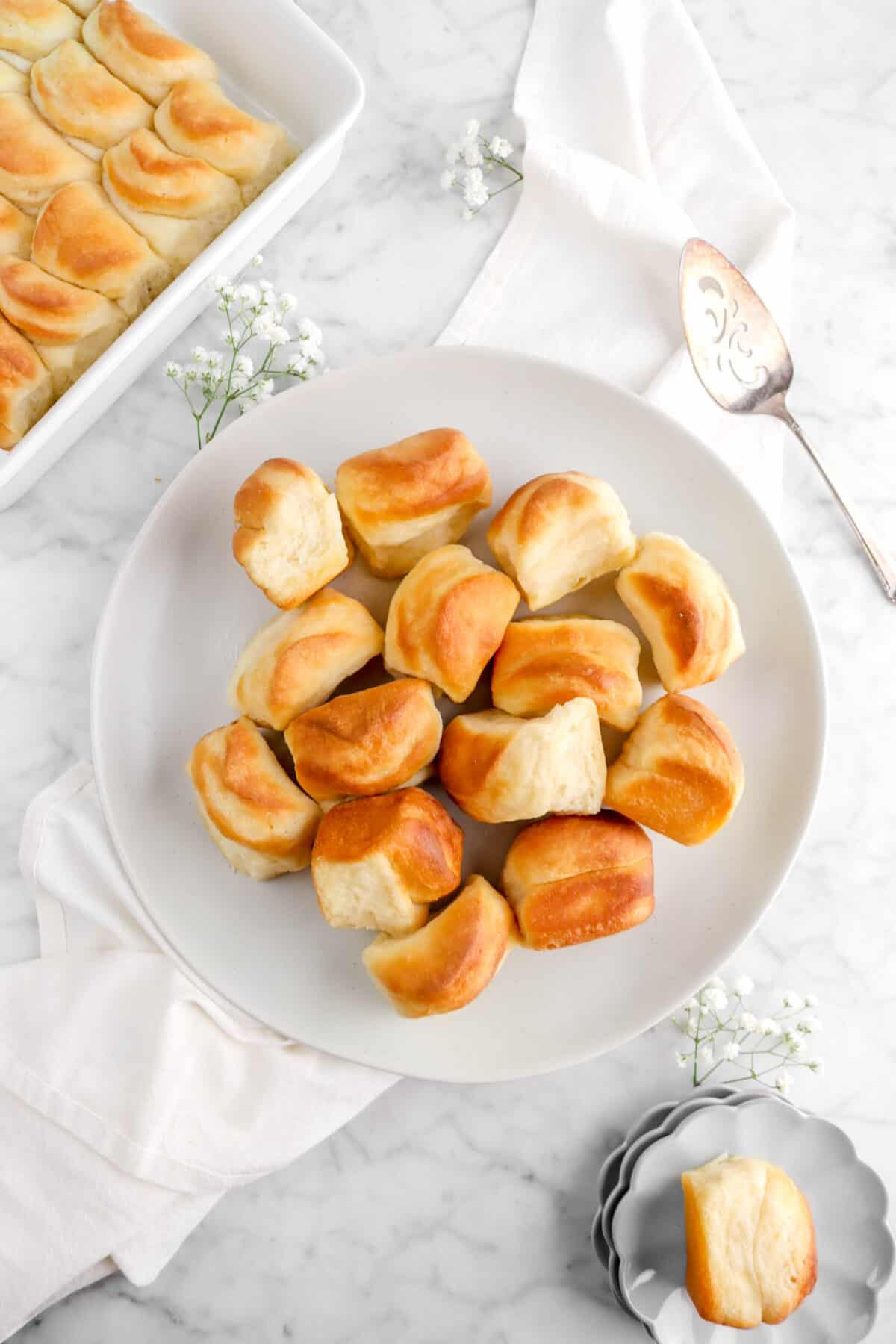 overhead shot of rolls on off-white plate with flowers, white napkin, roll on small grey plates, and casserole full of rolls