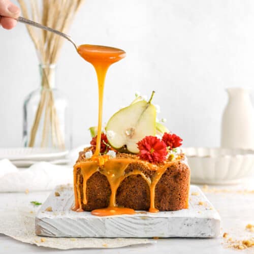 spoon dripping caramel onto loaf with flowers and sliced pear on top