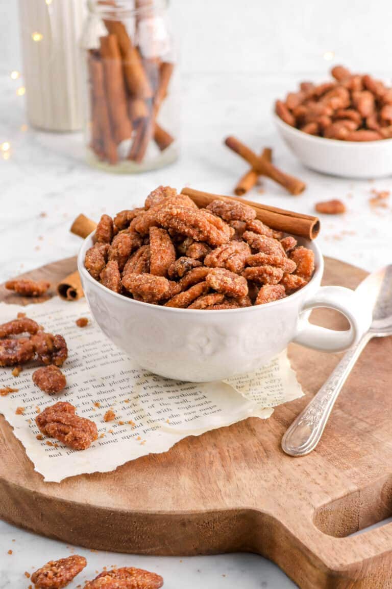 candied pecans in white mug with fairy lights, cinnamon sticks, and more pecans scattered around