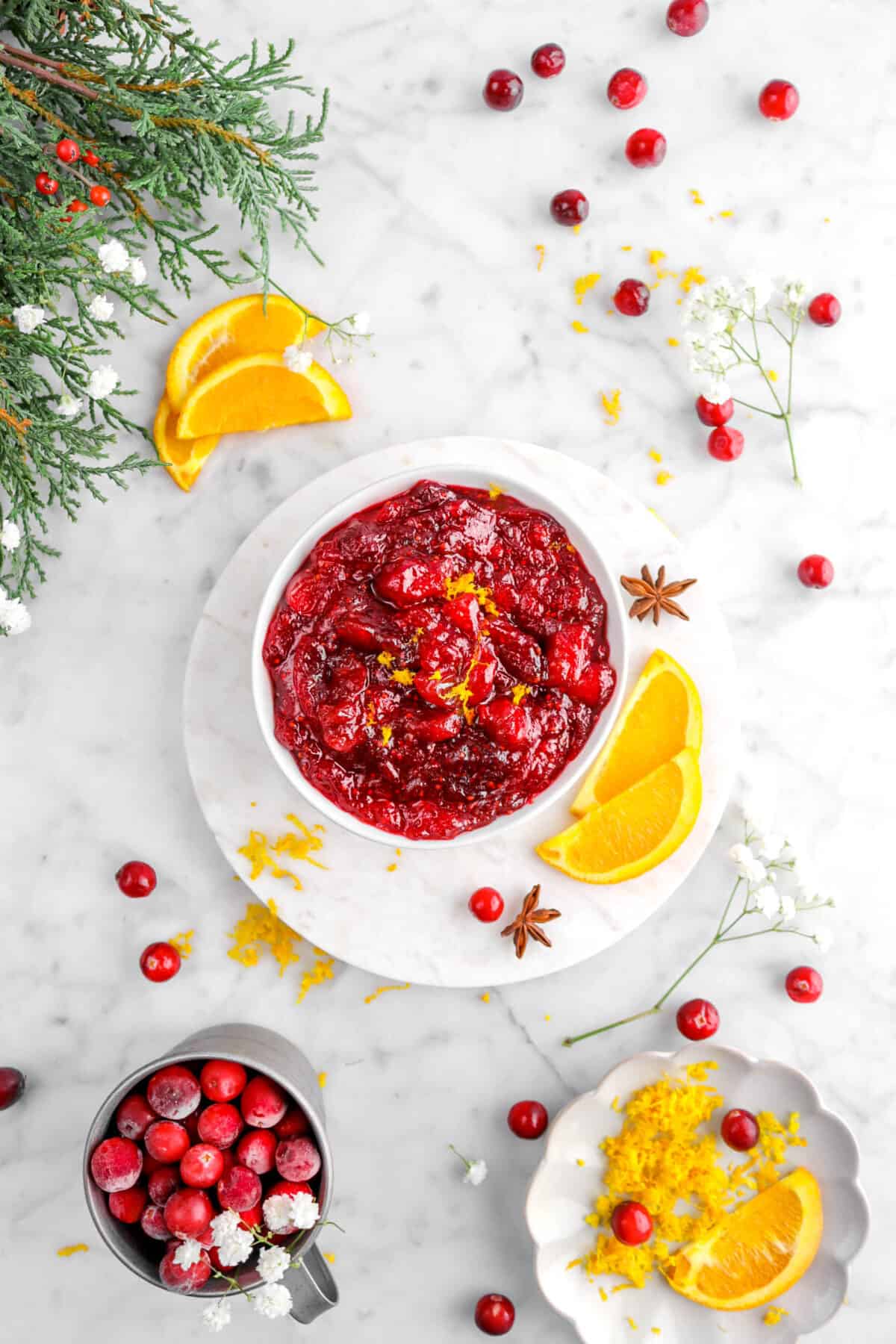 overhead shot of cranberry sauce in white bowl with orange zest, orange slices, two star anise pods, fresh cranberries, and greenery