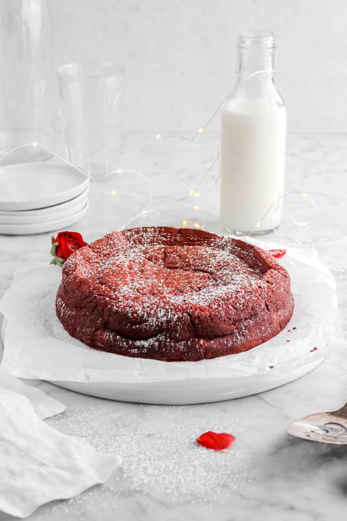 flourless red velvet cake on upside white palte with glass of milk, stacked plates, and powdered sugar