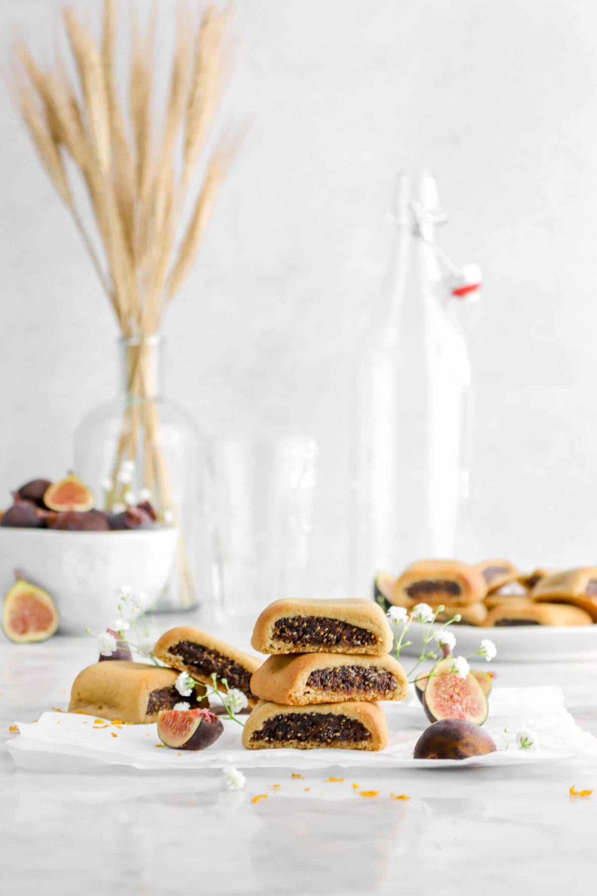 pulled back shot of fig newtons on parchment paper with fresh figs, jars, and a plate of fig newtons behind