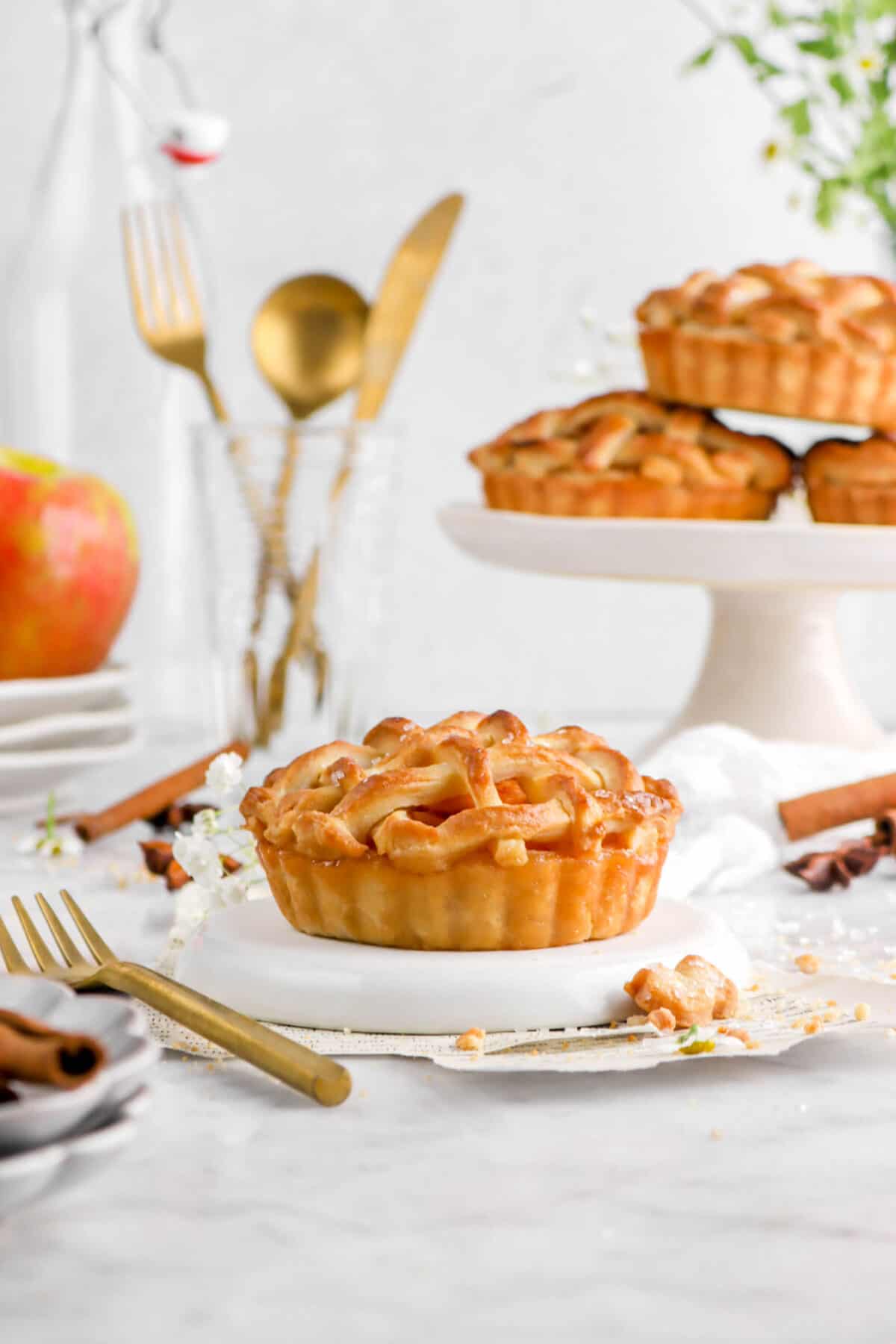 mini apple pie on upside down white plate with three pies behind, gold utensils, and an apple on stacked plates