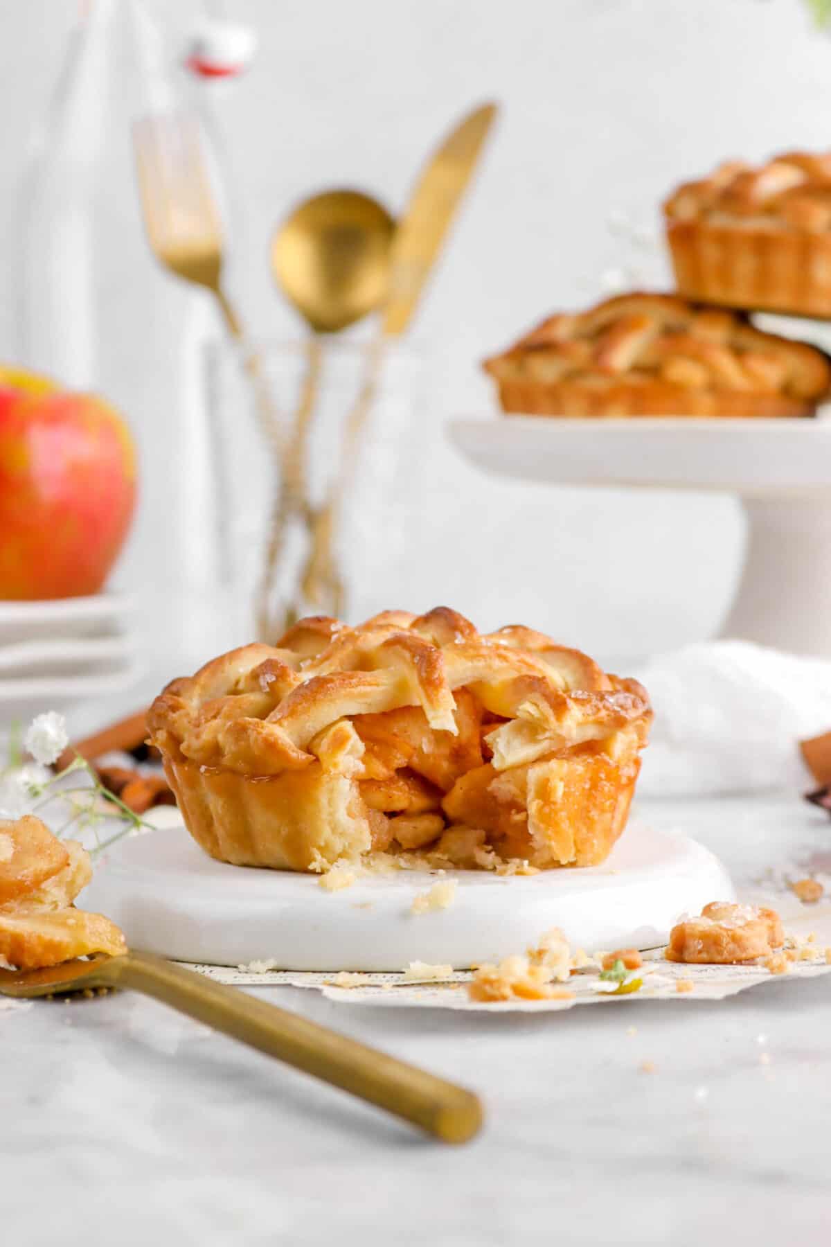 mini apple pie with bite missing on upside down white plate with gold utensils, an apple, and more pies