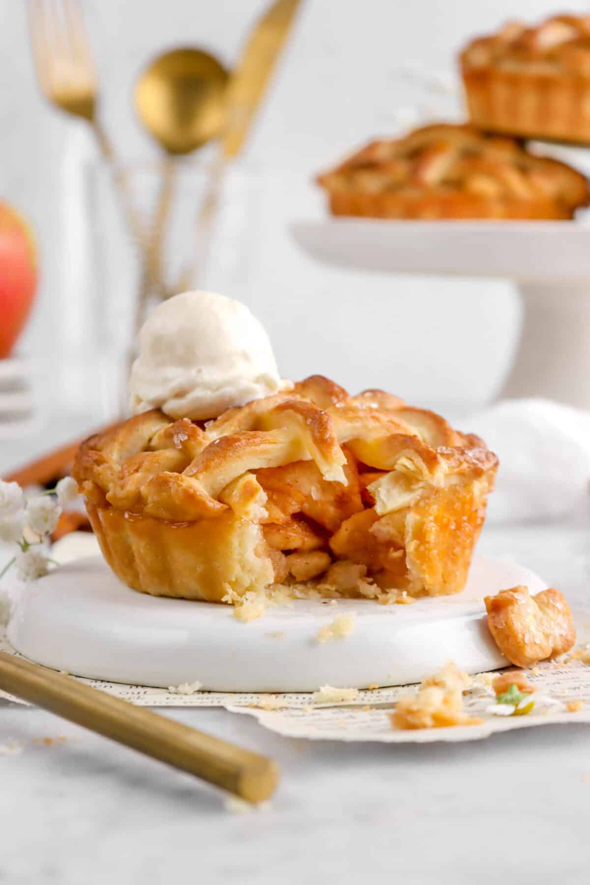 mini apple pie with scoop of vanilla ice cream on upside down white plate with gold utensils and two pies behind