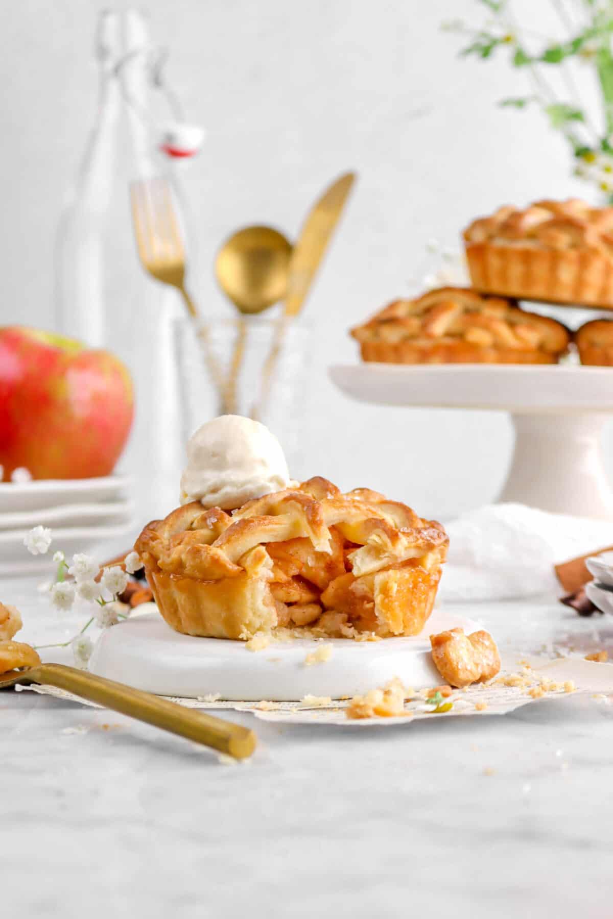 mini apple pie with scoop of ice cream, flowers, gold fork, an apple on stacked plates, and three pies behind