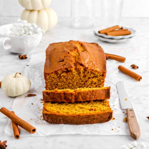 pumpkin bread on parchment paper with whole spices, two slices laying in front, a knife, flowers, and pumpkins