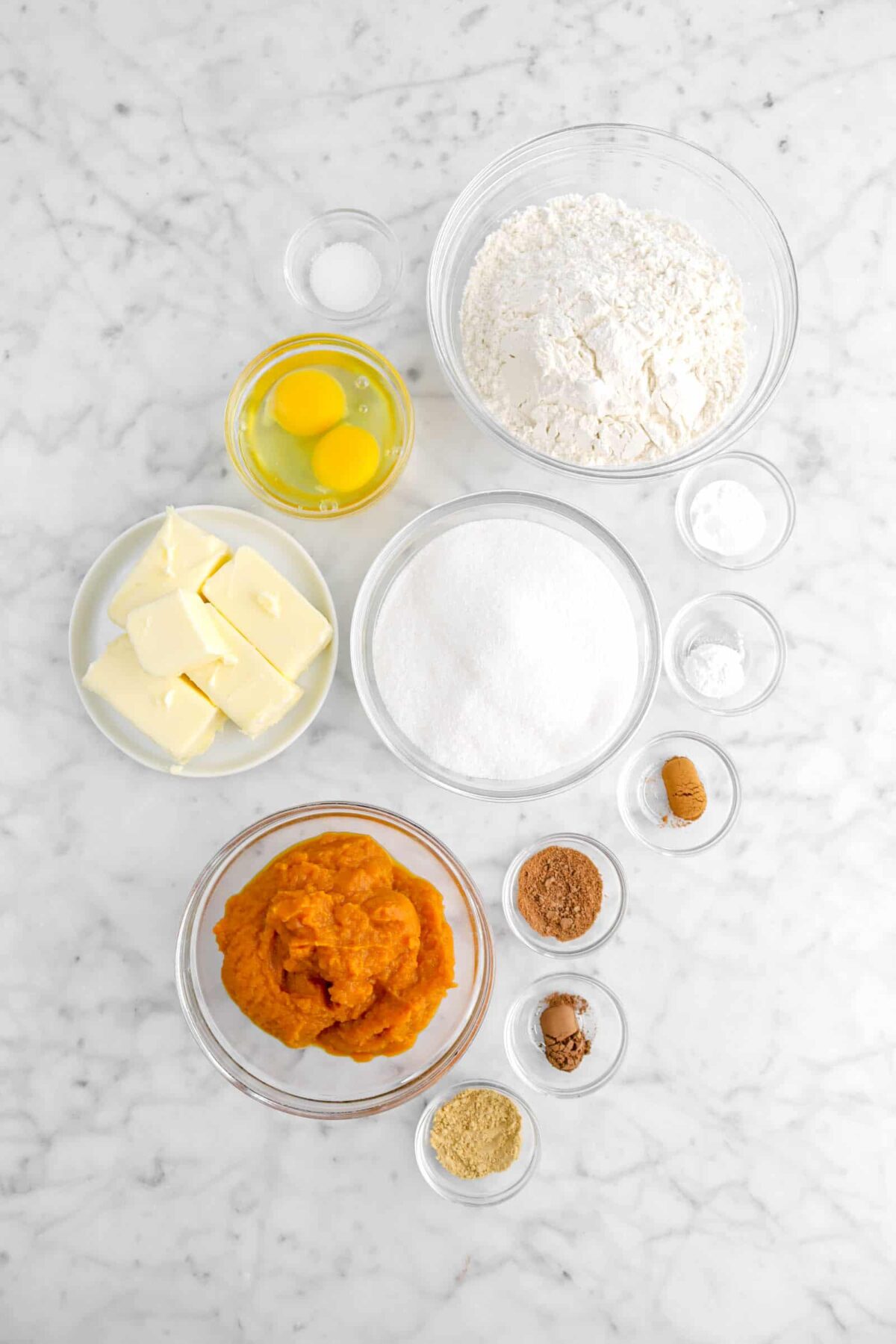 salt, flour, eggs, butter, sugar, baking soda, baking powder, cinnamon, nutmeg, cloves, ginger, and pumpkin puree on marble counter