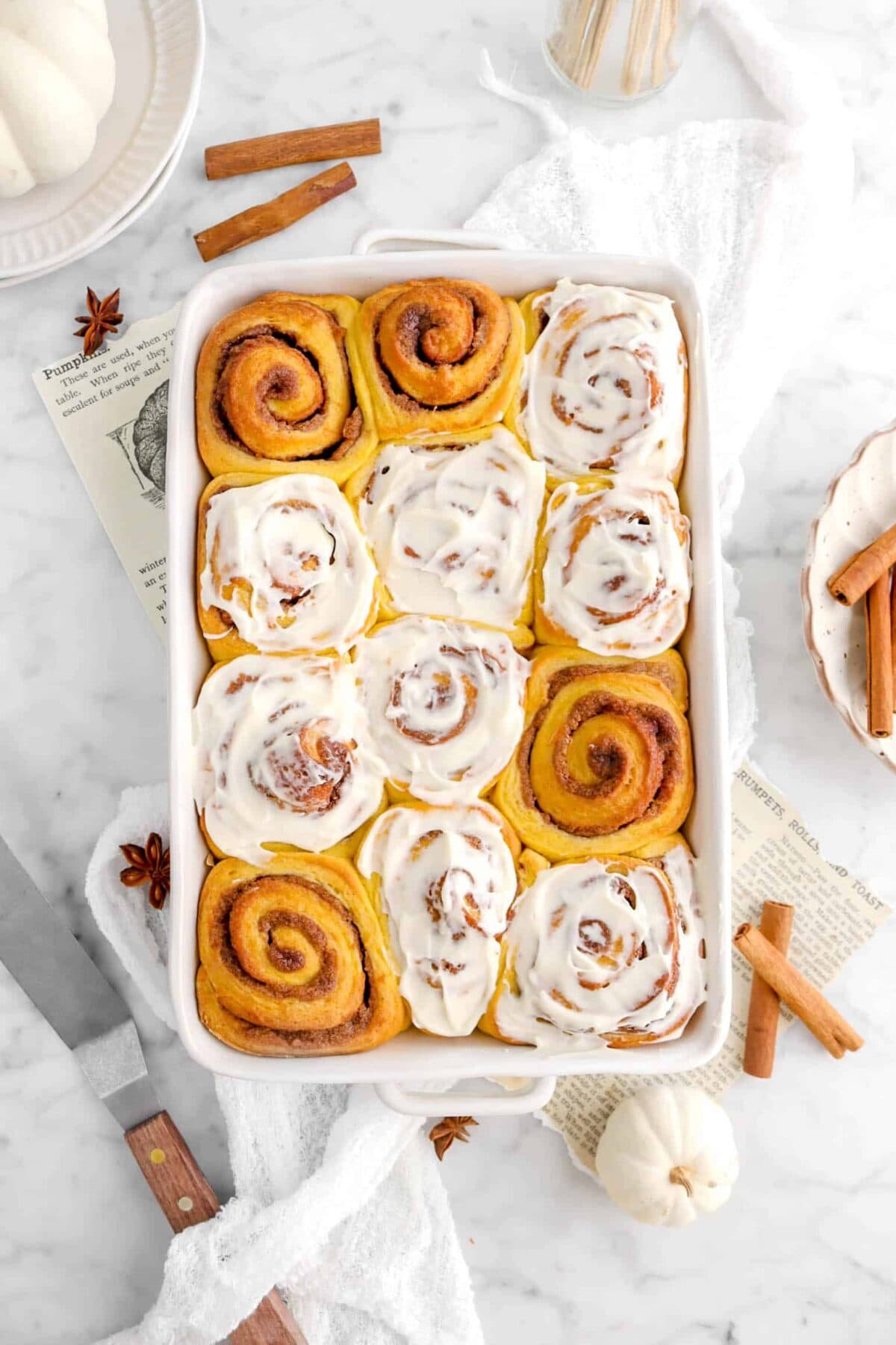 pumpkin cinnamon rolls with frosting, on white cheese cloth, and cinnamon sticks with white pumpkins