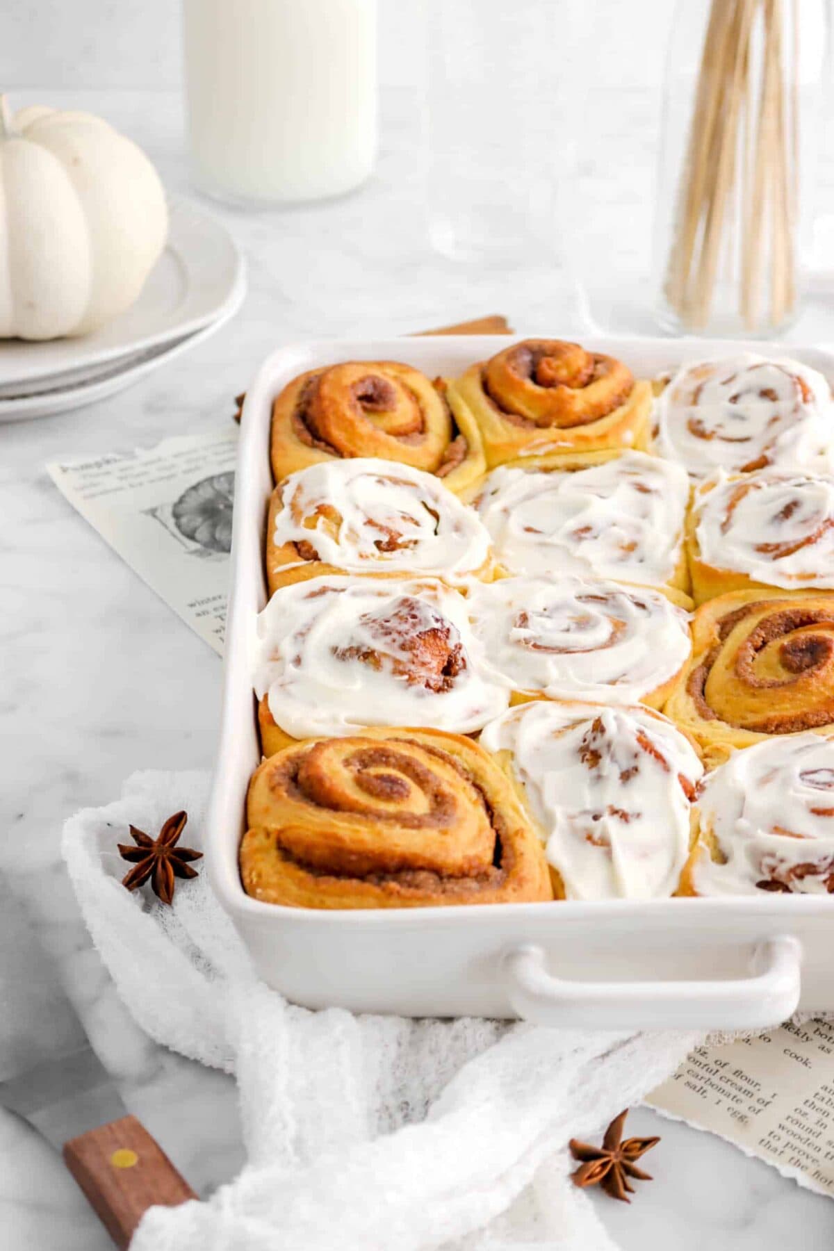 pumpkin cinnamon rolls in white casserole with white cheese cloth, plates, and star anise pods