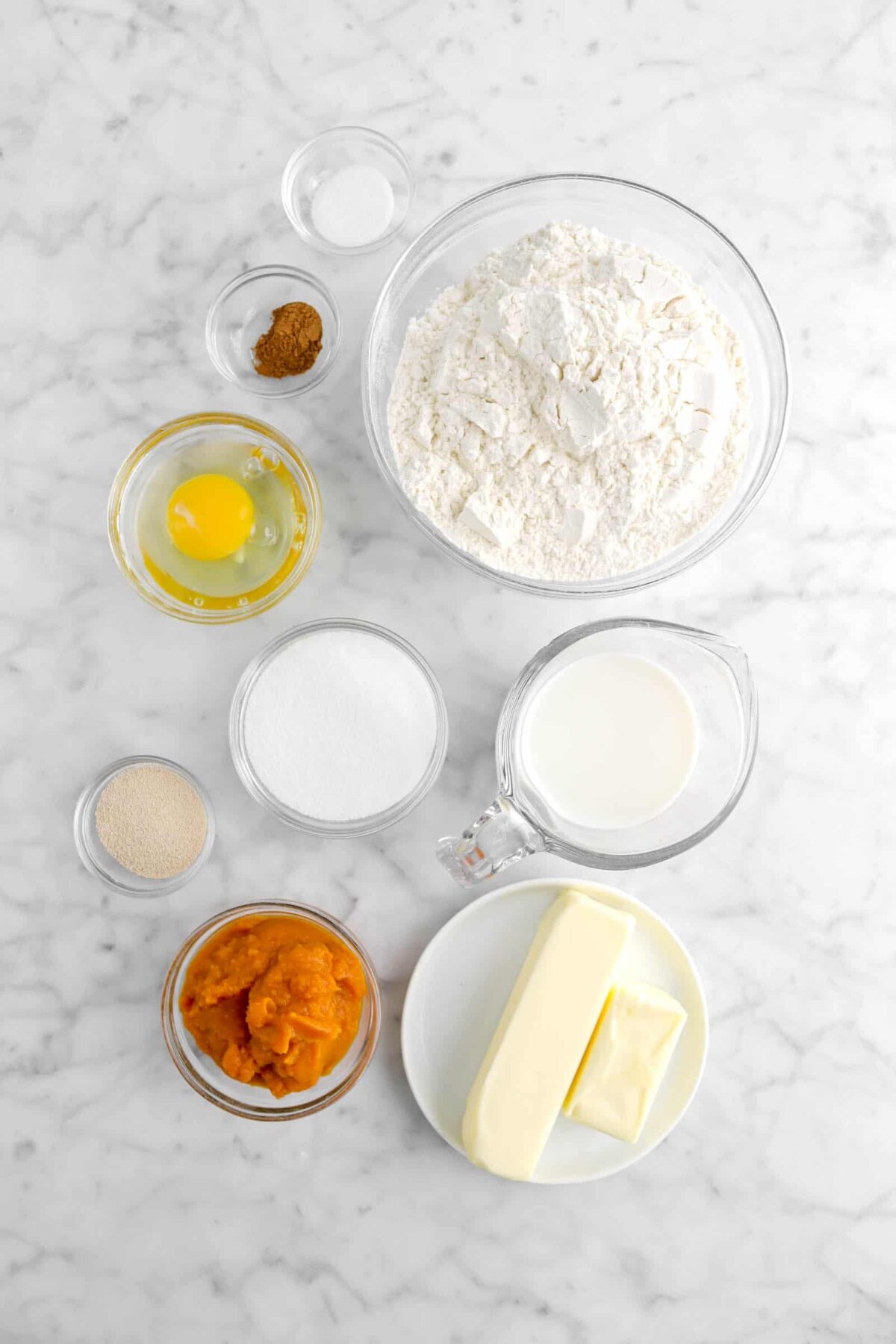 salt, pumpkin pie spice, flour, egg, sugar, milk, butter, pumpkin, and yeast on marble counter