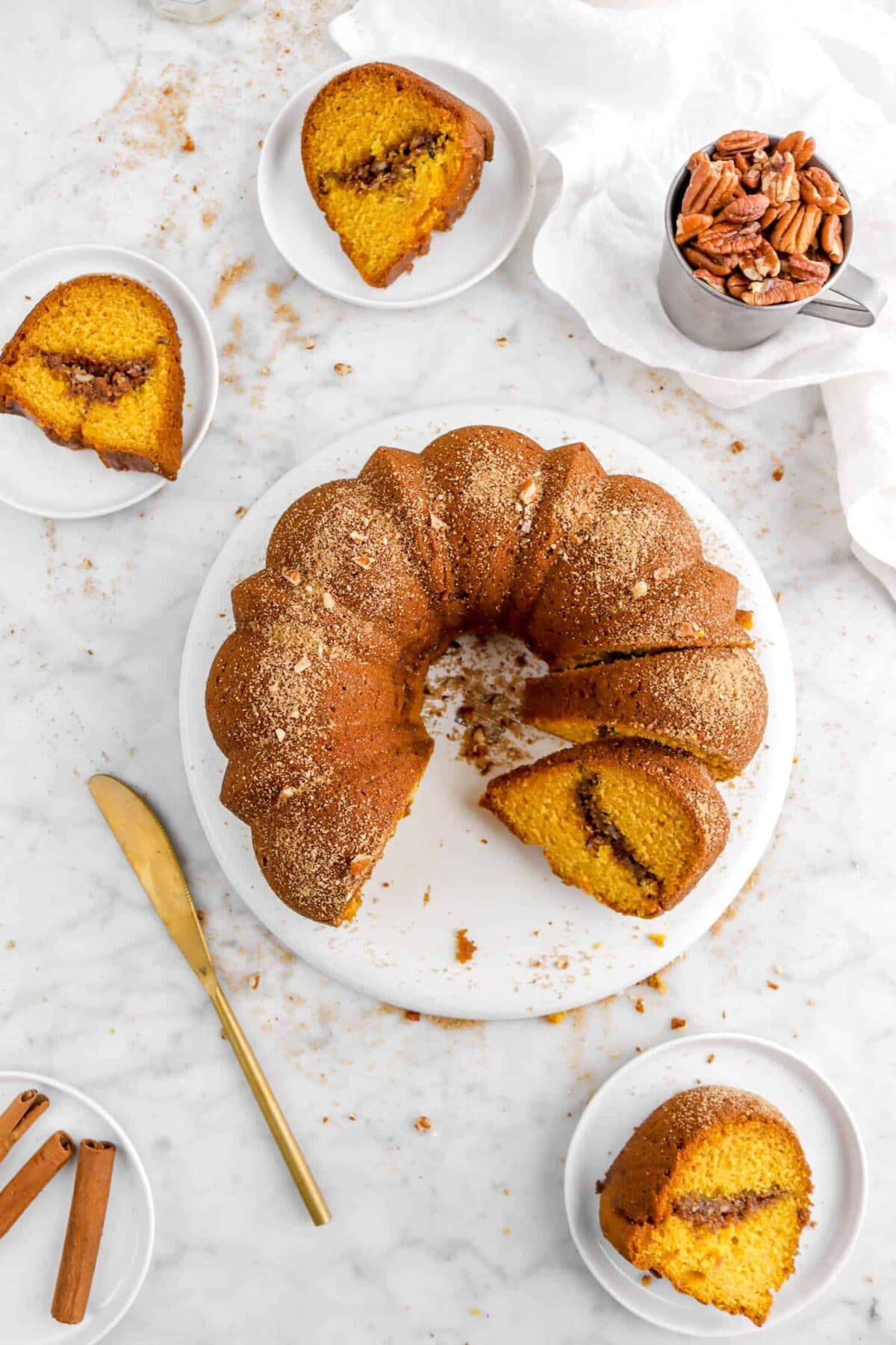 overhead shot of pumpkin coffee cake with gold knife, three plates around with slices of cake, and cup of pecans on white napkin