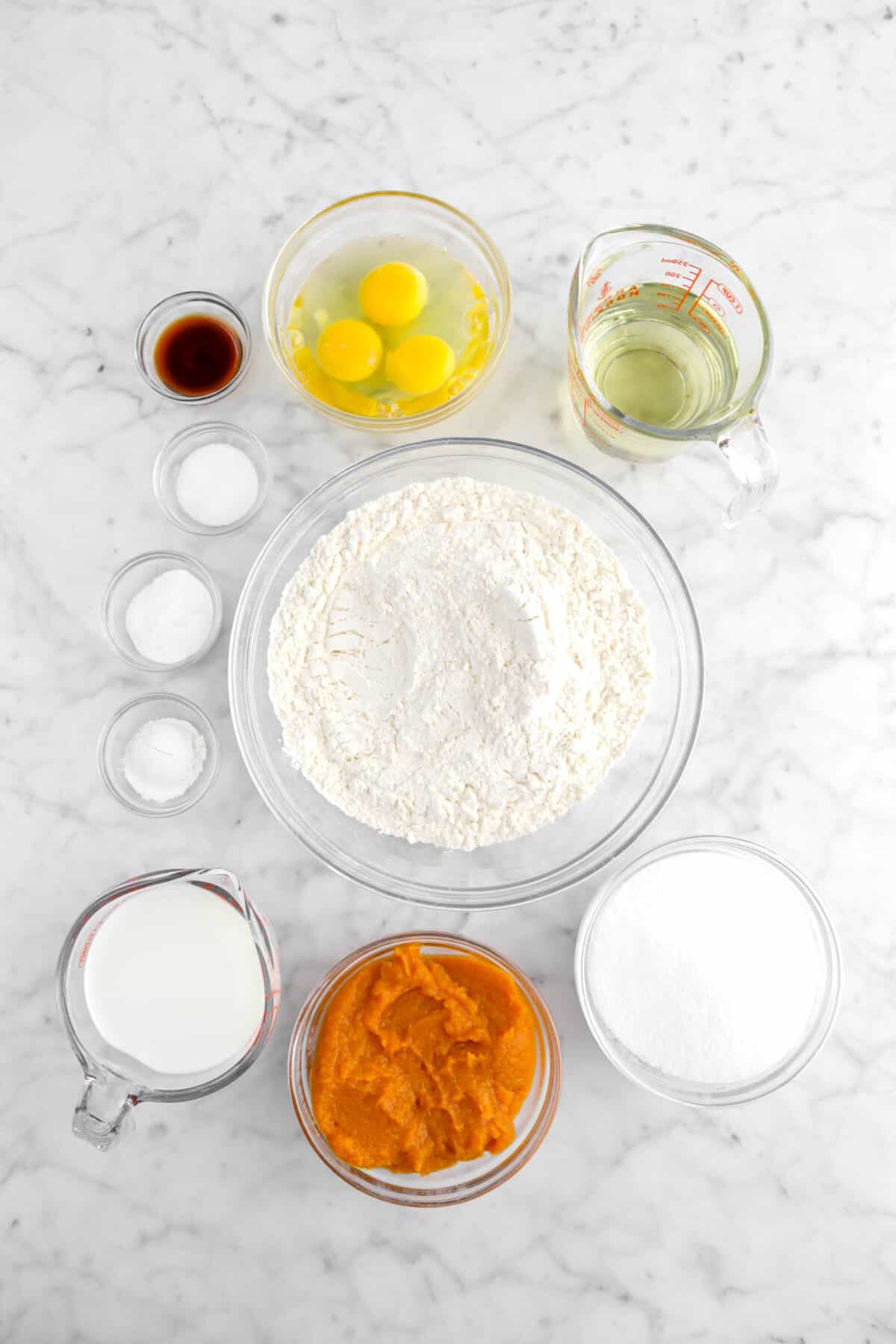 vanilla extract, eggs, vegetable oil, salt, baking soda, baking powder, flour, milk, pumpkin puree, and sugar in glass bowls on marble counter