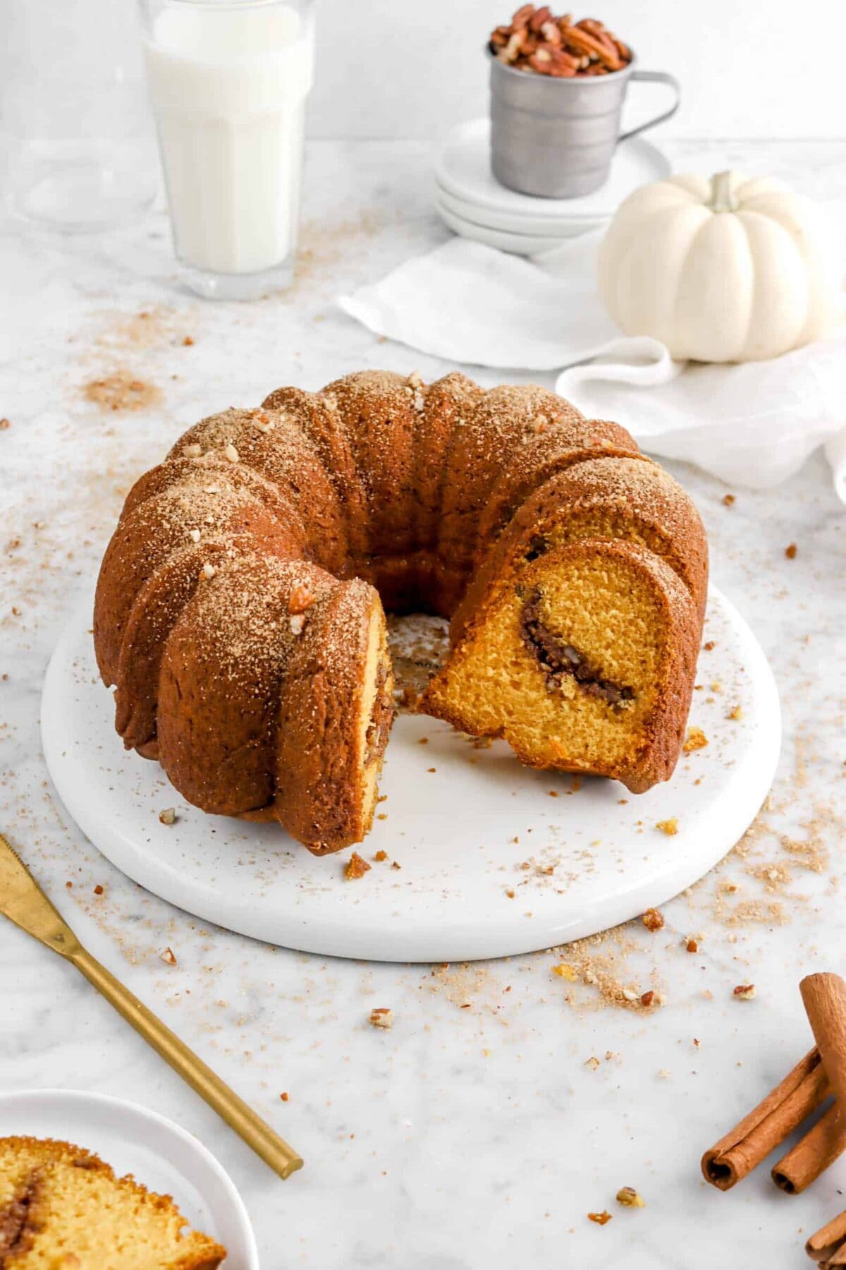 pumpkin bundt cake with glass of milk, cup of pecans, stacked plates, white napkin, and white pumpkin around