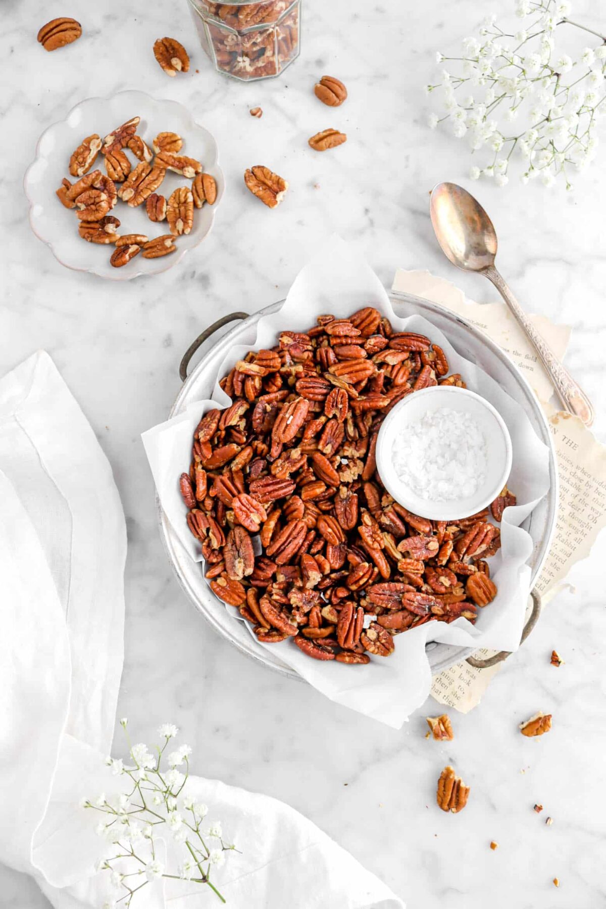 flat lay of roasted pecans in pan with bowl of salt, a spoon beside on top of book pages, flowers, and a white napkin