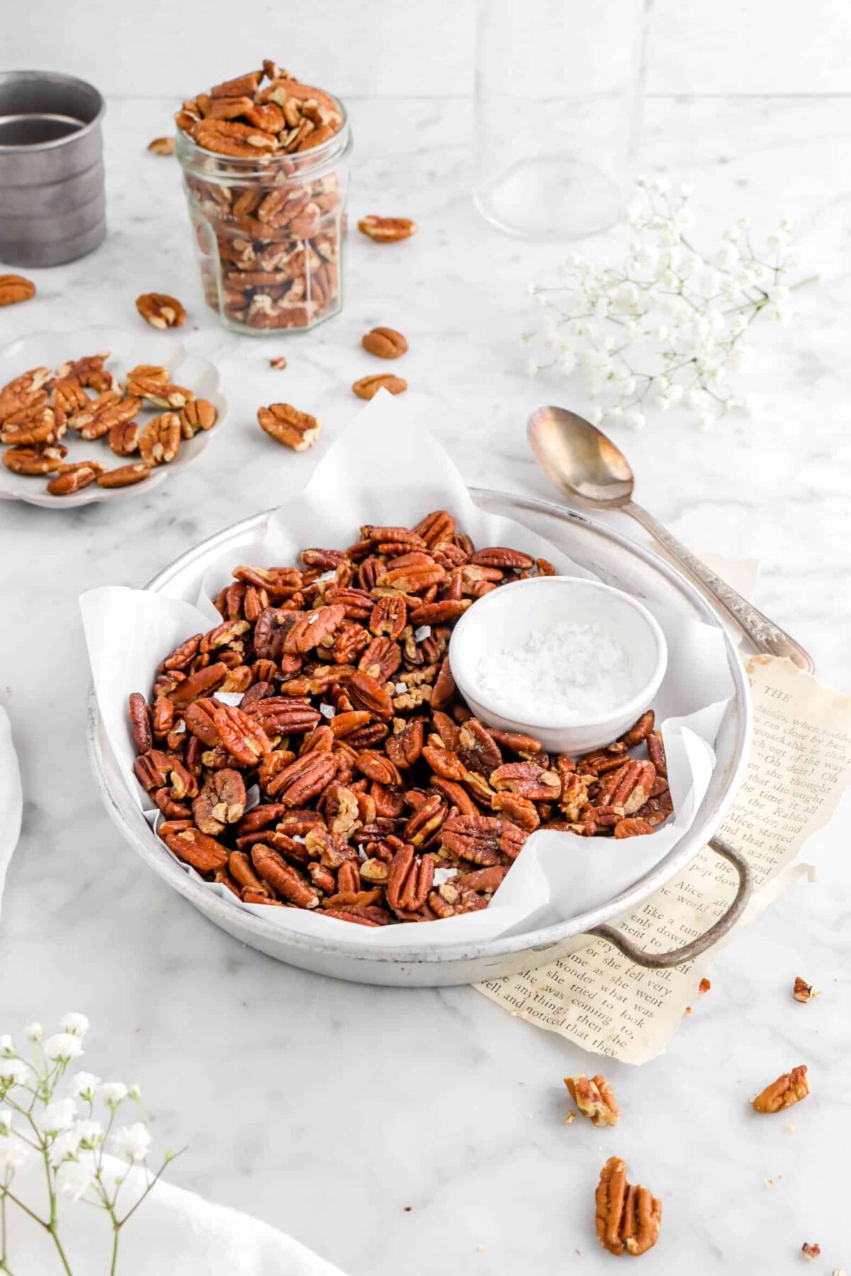 side shot of roasted pecans in old cake pan with flaked salt on top, bowl of salt, a spoon, more pecans around, and flowers