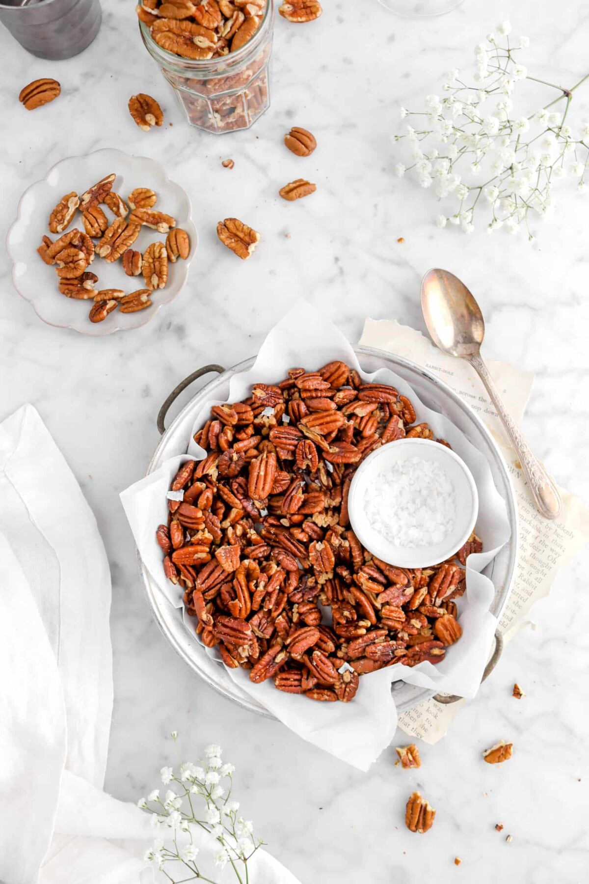 pecans in round pan with salt, a spoon beisde on top of book pages, flowers, plate and jarful of pecans, and a white napkin