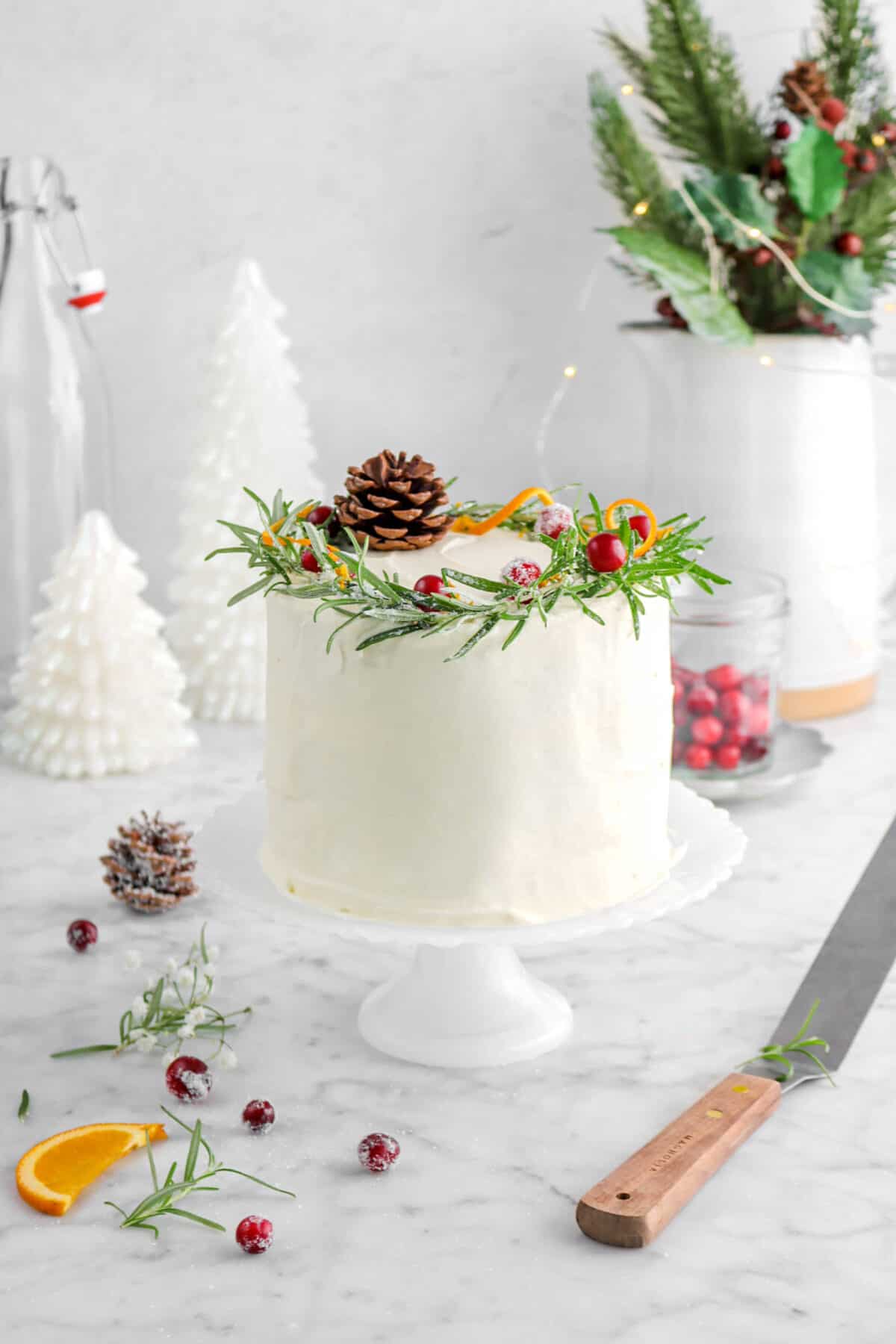 frosted cake on cake plate with rosemary, pine cone, cranberries, and orange twists on top with more around, white christmas trees behind, and christmas greenery with fairy lights