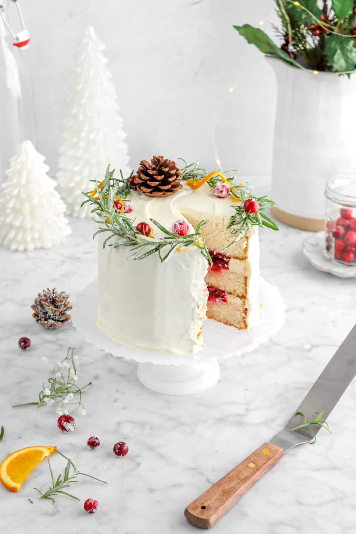 white cake with cranberry filling on cake plate with slice missing, offset spatula beside, jar of cranberries, flowers, rosemary sprigs around, and two white christmas trees behind
