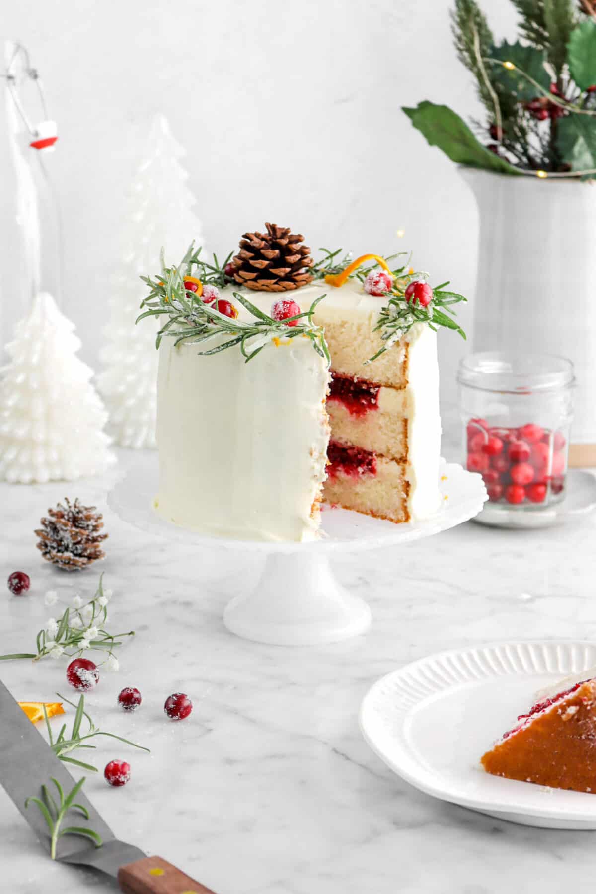 white cake on cake stand with offset spatula, cranberries, flowers, and pine cone beside, cake slice on white plate , with jar of cranberries and white christmas trees behind