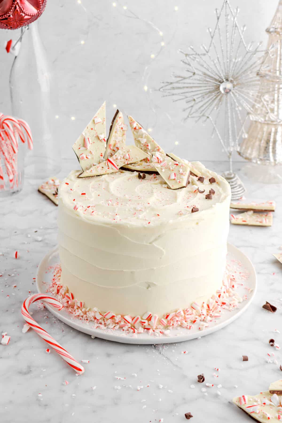peppermint cake with peppermint frosting on white plate with candy cane beside, peppermint bark on top, and lights behind