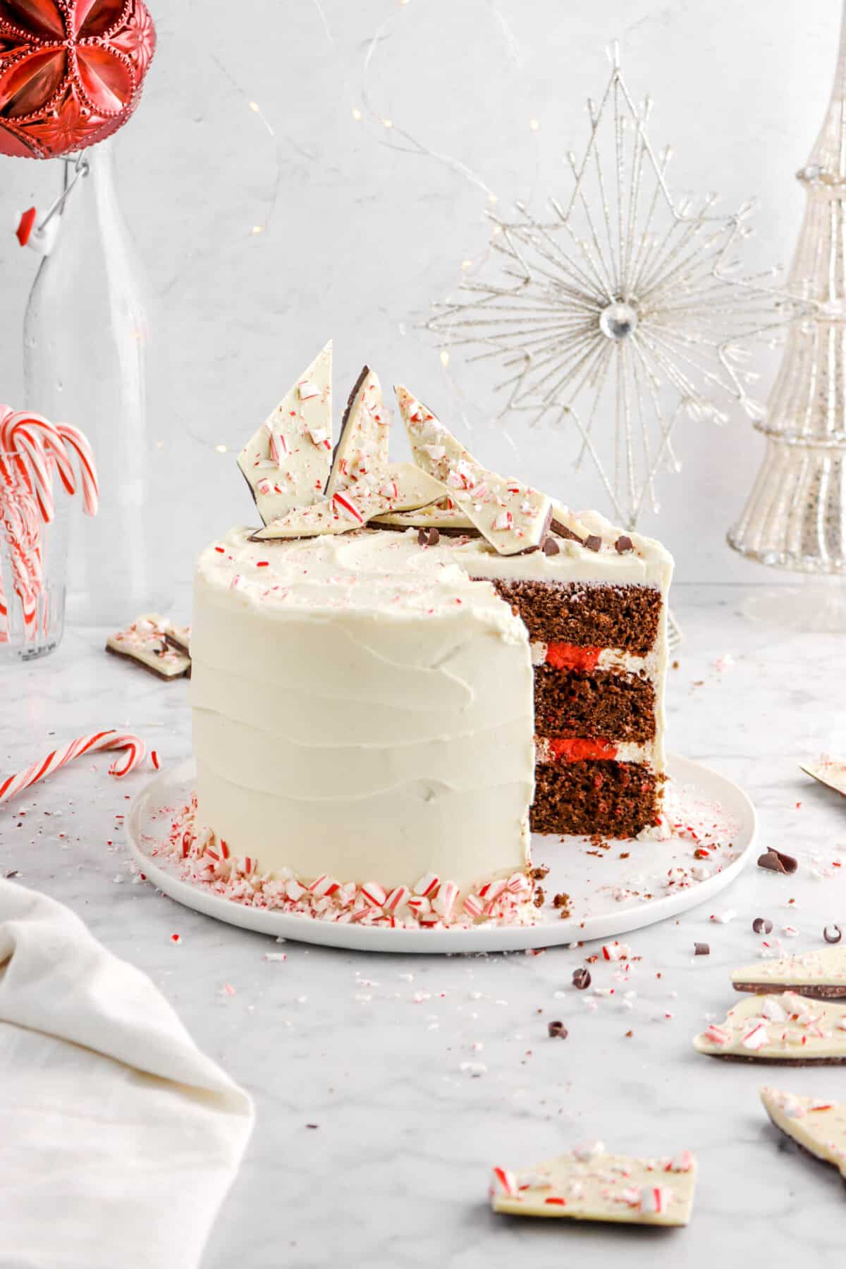 chocolate peppermint cake with slices taken out of cake on white plate with peppermint bark on top, decorative star, mercury glass tree, and red ornament